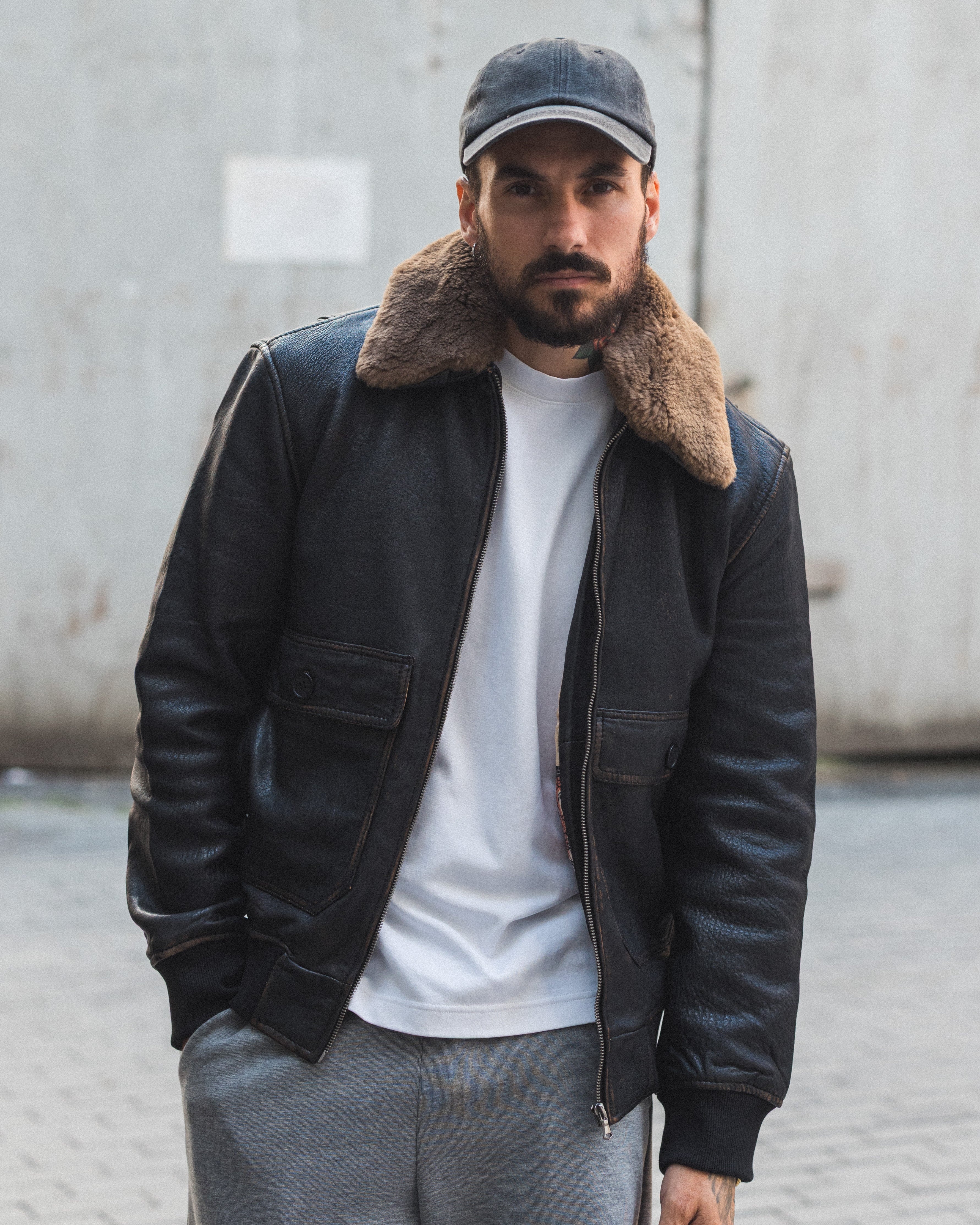 Man wearing a black leather jacket with fur collar, white t-shirt, and gray pants standing against a concrete wall.